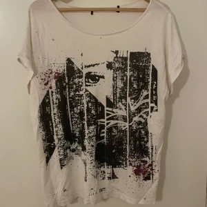 Vit t-shirt med grafiskt tryck - Vit t-shirt med ett stort grafiskt tryck i svart och lila framtill, föreställande ett abstrakt ansikte. T-shirten har rund halsringning och korta ärmar. Passformen är lös och avslappnad, perfekt för en cool och modern stil.