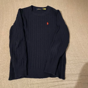 Mörkblå stickad tröja från Polo Ralph Lauren - Mörkblå stickad tröja från Polo Ralph Lauren med rund halsringning och klassisk röd broderad logga på bröstet. Tröjan har lång ärm och ribbade muddar vid ärmslut och nederkant. 