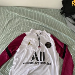 PSG fotbollströja med guldlogga - Säljer en PSG fotbollströja från Nike med vita och vinröda färger. Tröjan har guldiga detaljer, svart klubbmärke och sponsortryck på bröstet. Dragkedja vid halsen och långärmad modell. Perfekt för fans av Paris Saint-Germain.