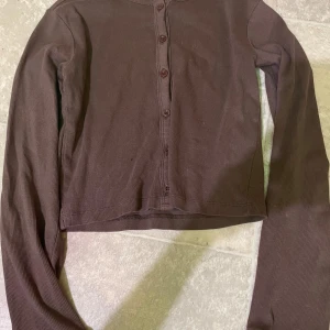 Brun ribbad kofta med knappar - Säljer en brun ribbad kofta med långa ärmar och knäppning framtill. Modellen är croppad och passar perfekt till lager-på-lager outfits. Enkel och stilren design som funkar till mycket.