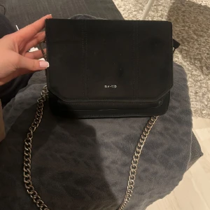 Svart axelväska från NA-KD med kedjerem - Snygg svart axelväska från NA-KD med silverfärgad kedjerem och dragkedja inuti. Den är i mocka. Köpt för 499 och säljer för 120kr🤍Man får även med ett tjockt axelband som satt på orginalväskan när den köptes! 