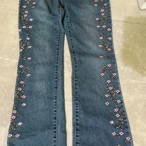 Blommiga jeans med broderi - Snygga blå jeans med broderade rosa blommor längs benen. Byxorna har en rak passform och klassisk jeanslook, perfekt för dig som vill ha något unikt i garderoben. Blommorna ger en söt och trendig touch till outfiten.