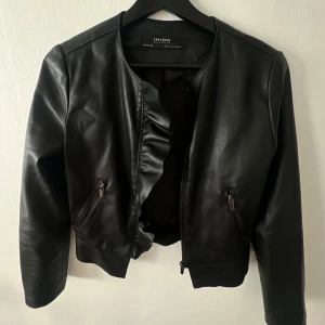 Svart skinnjacka med volang från Zara  - Snygg svart skinnjacka från Zara med volangdetalj längs dragkedjan och nertill. Jackan har rund halsringning, ganska långa ärmar för att vara XS och två fickor med dragkedja framtill. Den är lite sliten i kragen samt har ett ganska hål i innertyget vid ena armen (se bild). Det är sömmen som spruckit så det bör vara ganska enkelt att sy ihop. Inga skavanker på utsidan. 