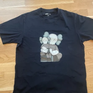 Svart t-shirt med KAWS-motiv från Uniqlo - Svart t-shirt från Uniqlo med tryck av KAWS-figurer på bröstet. Klassisk passform och rund halsringning. Perfekt för dig som gillar streetwear och snygga prints.