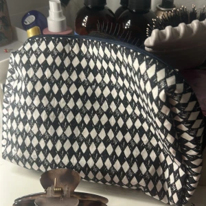 Svartvit necessär med diamantmönstertygg - Säljer en snygg necessär med svartvitt diamantmönster på utsidan och rödprickigt foder på insidan. Väskan har dragkedja upptill och är perfekt för att organisera smink eller småprylar. Praktisk och stilren accessoar till badrummet eller resan.