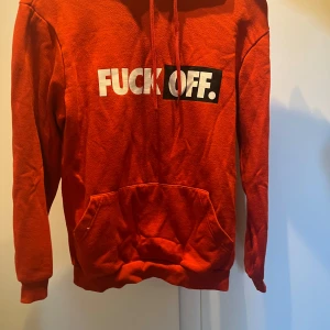 Röd hoodie med tryck - Säljer en röd hoodie med vit och svart text på bröstet. Tröjan har huva med dragsko och en stor magficka framtill. Perfekt för dig som vill sticka ut med ett kaxigt statement.