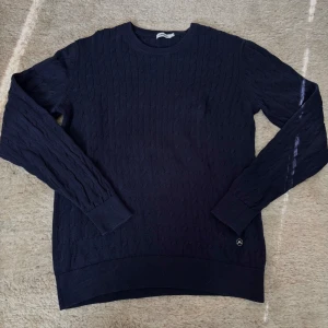 Mörkblå stickad tröja från J.Lindeberg - Säljer en mörkblå stickad tröja från J.Lindeberg. Tröjan har rund halsringning, diskret kabelstickat mönster och långa ärmar. Perfekt för lager-på-lager och stilrena outfits.