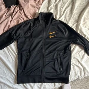 Svart zip från Nike med guldiga detaljer - Säljer en svart Nike ziptröja i storlek M med guldiga Swoosh-loggor på bröstet och textdetaljer på ryggen. Tröjan har dragkedja framtill och två fickor med dragkedja. Perfekt för sportiga tillfällen.
