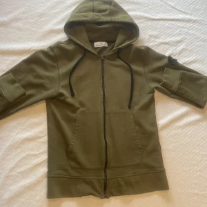 Olivgrön hoodie från Stone Island - Säljer en olivgrön hoodie från Stone Island med dragkedja och huva. Jackan har svarta snören, två stora fickor fram och den klassiska Stone Island-loggan på ärmen. Perfekt för en avslappnad och stilren look.