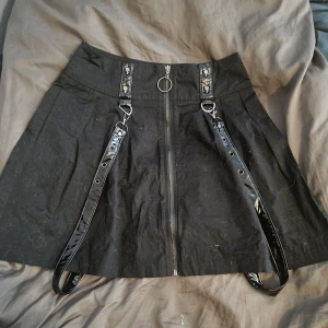 Svart kjol med dragkedja och hängslen från Banned Alternative - Cool svart kjol med silvrig dragkedja framtill och avtagbara hängslen med döskalle nitar. Kjolen har hög midja och detaljer i blankt material. Perfekt för dig som gillar alternativ stil.