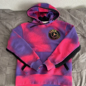 Paris Saint-Germain x Jordan hoodie i lila och rosa - Säljer en hoodie från Paris Saint-Germain x Jordan i lila och rosa med svart logga på bröstet och patch nertill. Nypris 1600kr