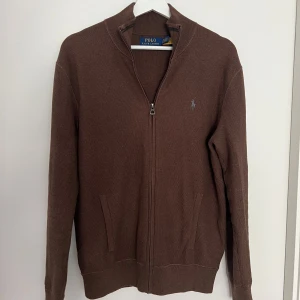 Brun stickad kofta från Polo Ralph Lauren - Säljer en brun stickad kofta från Polo Ralph Lauren med dragkedja framtill och två fickor. Klassisk design med broderad logga på bröstet och ribbade muddar vid ärmslut och nederkant. Perfekt för lager-på-lager. Nypris 2700kr.