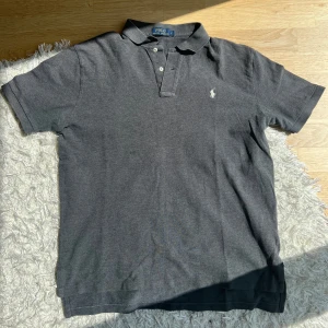 Grå pikétröja från Polo Ralph Lauren - Säljer en klassisk mörkgrå pikétröja från Polo Ralph Lauren med vit broderad logga på bröstet. Tröjan har korta ärmar, krage och knappar framtill. Perfekt för en stilren och avslappnad look. Hör av dig om du har fler frågor eller vill ha andra bilder.