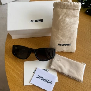 Svarta solglasögon från Jacquemus - Snygga svarta solglasögon från Jacquemus med rektangulära bågar och diskret Jacquemus-logga i guld på skalmen. Glasögonen har en stilren och modern design med breda skalmar och mörka linser. Kommer med originalförpackning, fodral och putsduk.