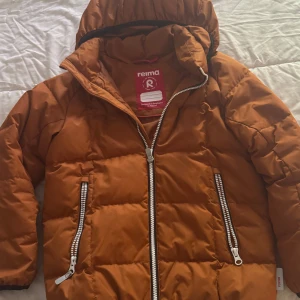 Orange pufferjacka från Reima - Säljer en orange pufferjacka från Reima med huva och två dragkedjeförsedda fickor. Jackan har långa ärmar och är perfekt för kyligare dagar. Snygg och praktisk design med tydliga detaljer. Den är lite förstörd vid armen men annars har den inga problem. Skön och håller barnen så varm! 