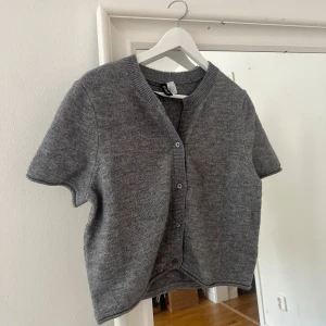 Grå stickad kofta med kort ärm - Grå stickad kofta från H&M med korta ärmar och knäppning framtill. Perfekt att slänga över en topp för en avslappnad look. Klassisk rund halsringning och mjukt material. Passar till sommarkvällar eller en kall sommardag till en långkjol eller liknande. 