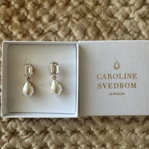 Pärlörhängen från Caroline Svedbom - Eleganta örhängen från Caroline Svedbom med rektangulär klar sten upptill och droppformad pärla nedtill. Guldfärgade detaljer och klassisk design, perfekta för att lyfta din look.
