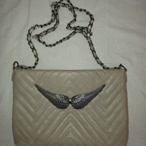 Beige axelväska med kedjerem och vingdetalj - Säljer en beige axelväska med quiltat mönster och snygg kedjerem. Väskan har en unik detalj framtill med silvriga vingar. Perfekt för dig som vill ha en stilren men ändå speciell accessoar.