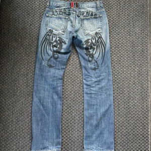 Angel Devil Jeans - Jeans från Angel Devil med text och tryck bak. Har även rhinestones på bakfickorna. Straight fit. Midjemått: 42cm Ytterben: 107cm Innerben: 84cm Benöppning: 21cm