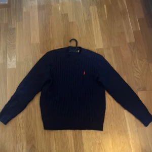 Mörkblå stickad tröja från Polo Ralph Lauren - Säljer en mörkblå stickad tröja från Polo Ralph Lauren med klassiskt röd broderad logga på bröstet. Tröjan har rund halsringning och långa ärmar, perfekt för kyligare dagar. Snygg och tidlös modell som passar till mycket. Har blivit lagad vid högra axeln men syns knappt när man har den på sig 