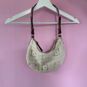Beige virkad axelväska med bruna detaljer - Superfin beige axelväska med virkat mönster och bruna axelremmar. Väskan har dragkedja upptill och en unik boho-vibe. Perfekt för att lyfta vilken outfit som helst!