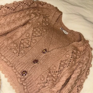 Rosa stickad kofta från Zara - Superfin rosa stickad kofta från Zara med dekorativa blommiga knappar och strukturstickat mönster. Koftan har lång ärm och v-ringning, perfekt för lager på lager. Söt och trendig detalj till din garderob!