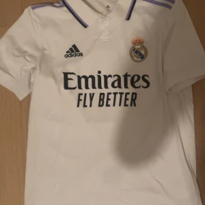 Real Madrid fotbollströja Benzema 9 - Säljer en vit Real Madrid matchtröja från Adidas med Benzema 9 på ryggen. Tröjan har korta ärmar, klubbmärke på bröstet och sponsortryck framtill. Klassisk design med lila detaljer vid krage och axlar. Pris kan diskuteras