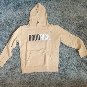 Beige hoodie från Hoodrich - Säljer en beige hoodie från Hoodrich i storlek medium. Tröjan har huva, magficka och tryckt logga 'HOODRICH.' på bröstet i svart och vitt. Perfekt för en avslappnad stil.