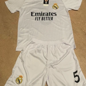 Real Madrid vit fotbollströja med shorts - Säljer en vit Real Madrid fotbollströja med tillhörande shorts. Tröjan har klubbmärke och sponsortryck på bröstet samt korta ärmar. Shortsen har klubbmärke och nummer 5. Perfekt för fotbollsträning eller match.
