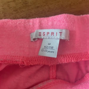 Rosa shorts från Esprit med dragsko - Säljer ett par knallrosa shorts från Esprit med elastisk midja och vit-rosa snörning. Shortsen har två fickor fram och ett tryckt blommönster med Esprit-logga på ena benet. Perfekta för varma dagar!