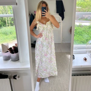 Blommig långklänning med volang - Säljer en långklänning med blommigt mönster i ljusa pastellfärger. Klänningen har volangdetaljer framtill. Perfekt för sommarens alla tillfällen!