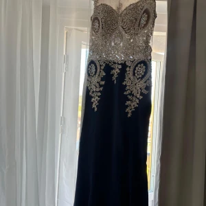 Mörkblå långklänning med glittriga detaljer - Elegant mörkblå långklänning med glittrig överdel och vackra paljettbroderier. Klänningen har ärmlös design och illusion neckline i mesh. Perfekt för bal eller fest. Snygg passform som framhäver figuren.