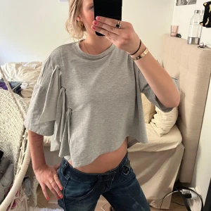 Trendig tröja! - Säljer en grå croppad t-shirt från Black Story i storlek XS. Tröjan har korta ärmar och coola knyt-detaljer på ena sidan som ger en unik look. Perfekt till jeans eller kjol för en avslappnad stil. Säljer då jag ej andvänder!