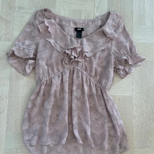 Ljusrosa blus  - Supersöt ljusrosa blus från H&M med diskret mönster och volangdetaljer vid halsringningen. Blusen har korta puffärmar och en lös passform, perfekt för sommaren. Hör av er vid frågor eller fler bilder😊