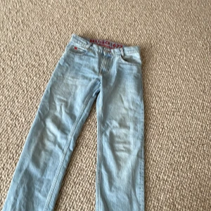 Ljusblå jeansbyxor från Hugo - Säljer ett par ljusblå jeansbyxor från Hugo med röd logga på insidan av linningen och liten röd detalj vid fickan. Klassisk femficksmodell med knapp och dragkedja. Perfekta för en avslappnad stil.
