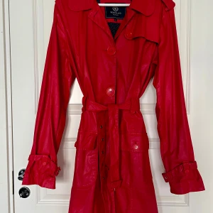 Röd trenchcoat från Henri Lloyd - Säljer en snygg röd trenchcoat i vaxat material från Henri Lloyd med klassisk krage, knappar och bälte i midjan. Jackan har långa ärmar och två fickor framtill. Perfekt för att sticka ut i mängden under vår och höst! Storlek 6 vilket mostvarar XXL men passar en XL. 
