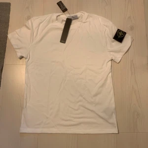 Vit t-shirt från Stone Island - Säljer en klassisk vit t-shirt från Stone Island med den ikoniska loggan på vänster ärm. T-shirten har rund hals och är tillverkad i mjuk bomull. Perfekt för en stilren och enkel look.