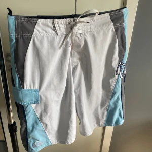 Quiksilver badshorts i blått och vitt - Säljer ett par snygga badshorts från Quiksilver i ljusblått, mörkblått, vitt och grått. Shortsen har snörning i midjan och stort broderat Quiksilver-tryck på sidan. Perfekta för stranden eller poolen! Riktigt snygga att ha på sig på sommaren. Säljer pga växt ur dem.