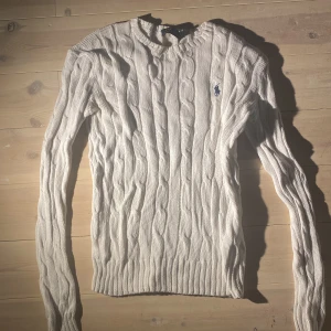 Vit kabelstickad tröja från Polo Ralph Lauren - Säljer en klassisk vit kabelstickad tröja från Polo Ralph Lauren med lång ärm och rund hals. Tröjan har den ikoniska blå loggan broderad på bröstet och är tillverkad i mjukt material. Perfekt för lager-på-lager under kyliga dagar. Köpt på plick för 700kr