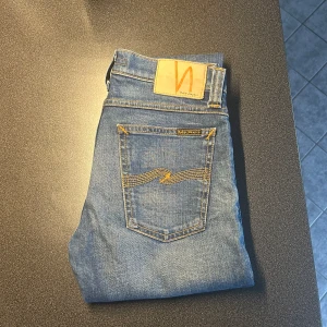 Blå jeans från Nudie Jeans - Säljer ett par klassiska blå jeans från Nudie Jeans med raka ben och snygga kontrastsömmar. Byxorna har fem fickor och Nudie-logga på bakfickan. Perfekta för dig som gillar stilrena och tidlösa jeans.
