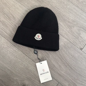 Svart ribbad mössa från Moncler - Svart ribbstickad mössa från Moncler med uppvikt kant och klassisk Moncler-logga framtill. Perfekt accessoar för kyliga dagar och enkel att matcha med olika outfits.