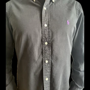 Svart skjorta från Ralph Lauren - Klassisk svart skjorta från Ralph Lauren med lila broderad logga på bröstet. Skjortan har knappar framtill och krage med knappdetaljer. Perfekt för en stilren look.