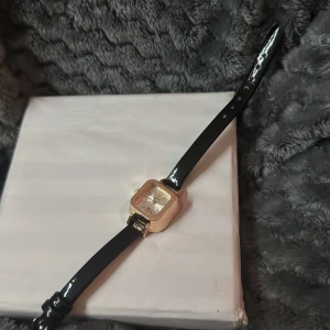 Klassisk damklocka med svart läderband och guldfärgad boett - Stilren klocka med fyrkantig guldfärgad boett och svart glansigt läderband. Urtavlan är ljus med tydliga markeringar och smala visare. Perfekt accessoar för dig som gillar en tidlös och elegant look.