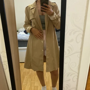 Beige trenchcoat med knappar - Säljer en klassisk beige trenchcoat med knappar framtill och krage. Jackan har långa ärmar och en stilren, rak passform som passar till många outfits. Perfekt för vår och höst.