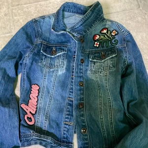 Blå jeansjacka med broderier - Säljer en blå jeansjacka med broderade detaljer. Jackan har knappar framtill, två bröstfickor och broderier med blommor och texten 'Amour' på framsidan. Perfekt för dig som vill ha en unik och personlig stil.