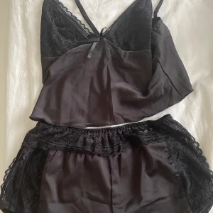 Svart satinlinne med spetsdetaljer - Säljer ett svart linne och shorts i satin med tunna axelband och spets upptill. Linnet har en liten rosett framtill och matchas med shorts i samma material och spets längs sidorna. Perfekt för en mysig kväll hemma.