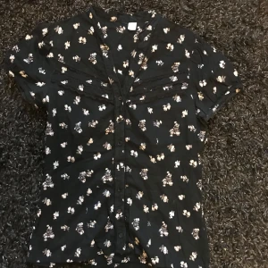 Svart blommig skjorta med kort ärm - Svart skjorta med små blommiga mönster i vitt och beige. Skjortan har korta ärmar och knappar framtill. Perfekt för en avslappnad och stilren look.