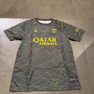 PSG fotbollströja med gult mönster - Säljer en svart PSG fotbollströja från Jordan med gula detaljer och grafiskt mönster. Tröjan har korta ärmar, rund hals och tryck med Qatar Airways på bröstet. Perfekt för dig som gillar snygga matchtröjor och vill sticka ut på planen.