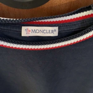 Mörkblå t-shirt från Moncler med röd och vit kant - Snygg mörkblå t-shirt från Moncler med rund halsringning som har röd och vit randig kant. Klassisk passform och Moncler-logga på ärmen. Perfekt för dig som gillar stilrena plagg med en sportig touch.