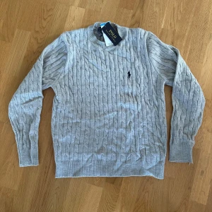 Grå knit  från Polo Ralph Lauren - Säljer en klassisk marinblå kabelstickad tröja från Polo Ralph Lauren. Använd 1-3 gånger innan jag sluta använda den. Väldigt fin och är i nyskick med inga skador eller liknande. (Säljer även en exakt likadan i marinblå , kolla profil ifall du vill köpa båda!)